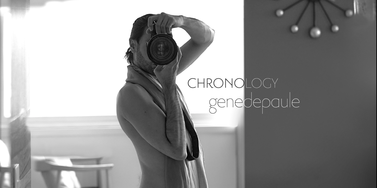 Black and white self portrait; SWCHPS wearing a shawl and holding the camera, photographing towards a mirror; behind him, the indoor setting of an apartment, almost empty except for and a wall mounted clock